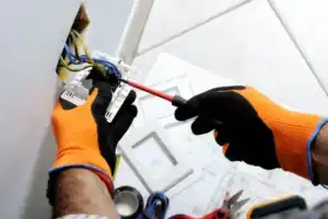 A Divine team member in orange gloves expertly repairs an electrical outlet, with exposed wires and tools neatly arranged nearby.